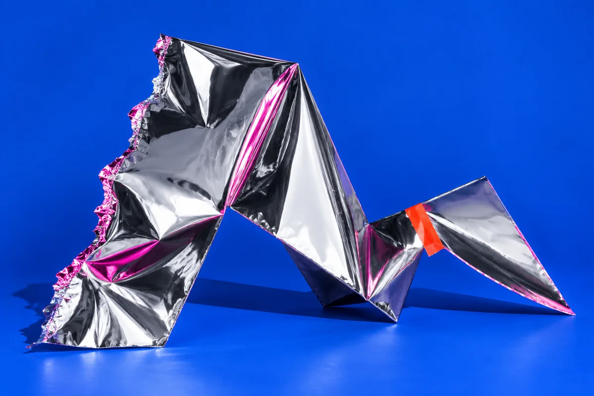 An unfinished chrome foil sculpture with a red tape repair on a cobalt blue surface.